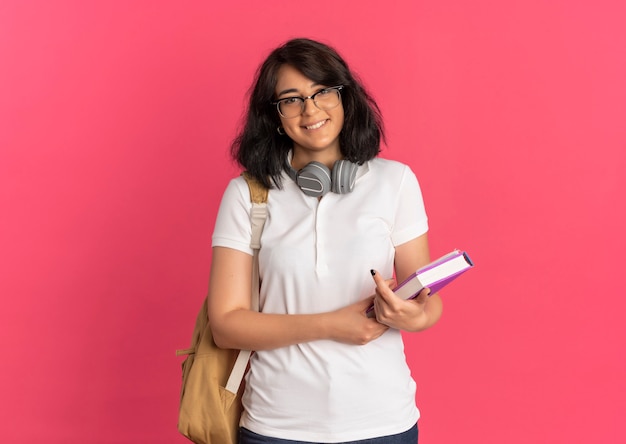 Foto grátis jovem sorridente, bonita caucasiana, usando óculos, fones de ouvido e bolsa traseira segurando um livro rosa com espaço de cópia