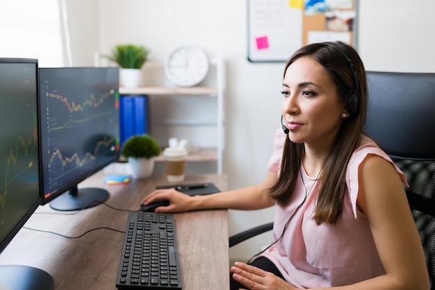 Foto grátis linda jovem usando um fone de ouvido para ligar para clientes enquanto olha para o monitor do computador e verifica o mercado de ações