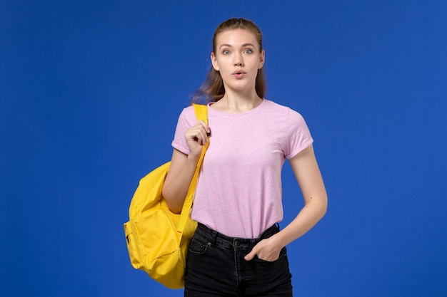 Foto grátis vista frontal de uma jovem mulher com uma camiseta rosa e uma mochila amarela em pé na parede azul