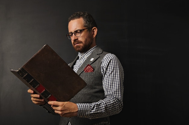 Kostenloses Foto ernsthafter männlicher lehrer in tweedweste und kariertem hemd, das ein großes altes buch an einer tafel liest