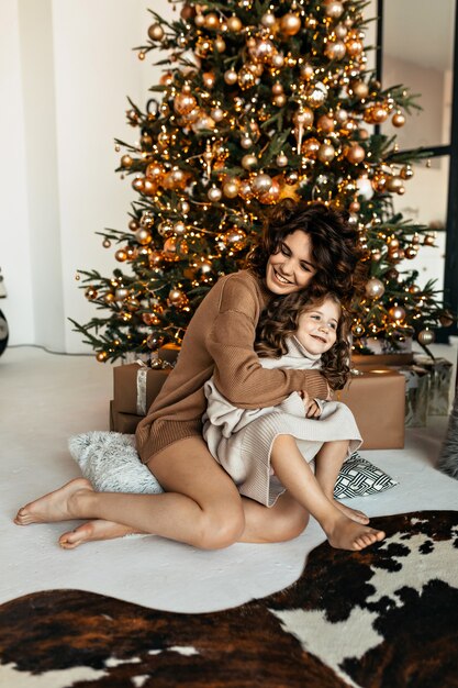 Glückliches Familienporträt der hübschen Mutter mit der Tochter gekleideten gestrickten Pullovern, die vor Weihnachtsbaum sitzen und Neujahr feiern
