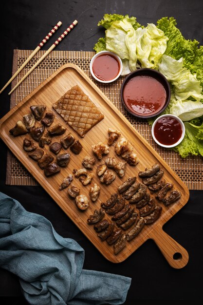 High Angle Shot eines Holztabletts mit gebratenem Fleisch, Kartoffeln, Gemüse und Sauce auf einem schwarzen Tisch