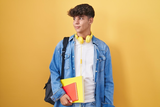 Kostenloses Foto hispanischer teenager, der einen studentenrucksack trägt und bücher hält, die lächelnd zur seite schauen und nachdenklich wegstarren.