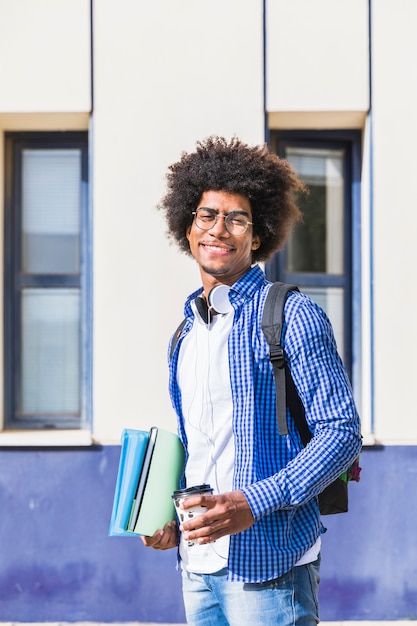Kostenloses Foto lächelnder tragender rucksack des jugendlichen männlichen studenten auf der schulter, die stapel der buch- und mitnehmerkaffeetasse steht am campus hält