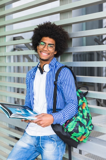 Kostenloses Foto porträt eines lächelnden afroen männlichen studenten, der in der hand das buch sich lehnt an der wand hält