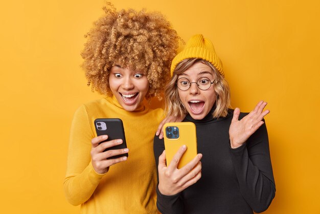 Technologie- und Freundschaftskonzept Verrückte positive Frauen benutzen Mobiltelefone starren auf den Bildschirm und reagieren auf erstaunliche Nachrichten, die in lässigen Rollkragenpullovern gekleidet sind, die isoliert über der gelben Wand stehen, bemerken etwas Unglaubliches