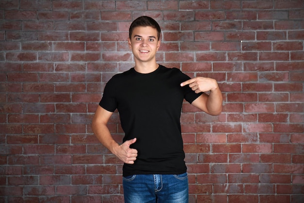 Foto apuesto joven en camiseta negra en blanco de pie contra la pared de ladrillo