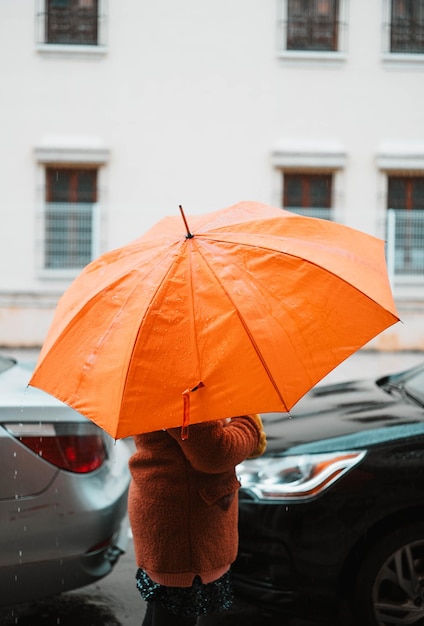 Foto chica con un paraguas naranja protegiéndose de la lluvia caminando entre los autos