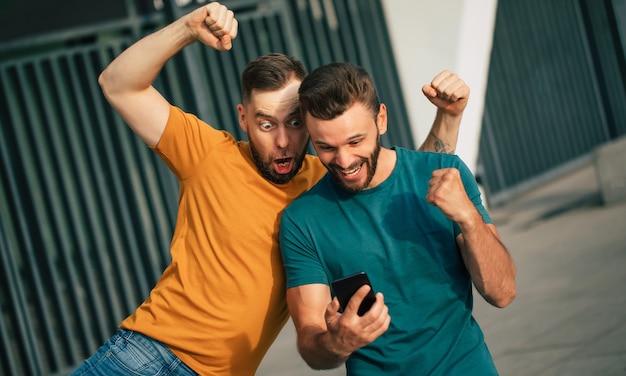 Foto dos amigos fanáticos emocionados felices en estado de ánimo de euforia después de ganar en una apuesta con un teléfono inteligente en la mano