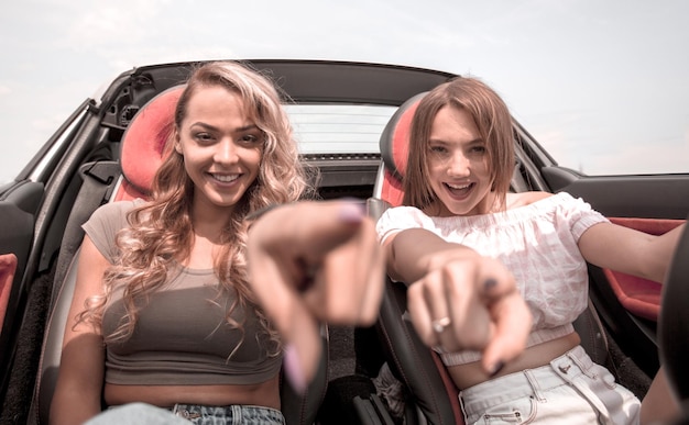 Foto dos mujeres jóvenes felices sentadas en un auto y señalándote