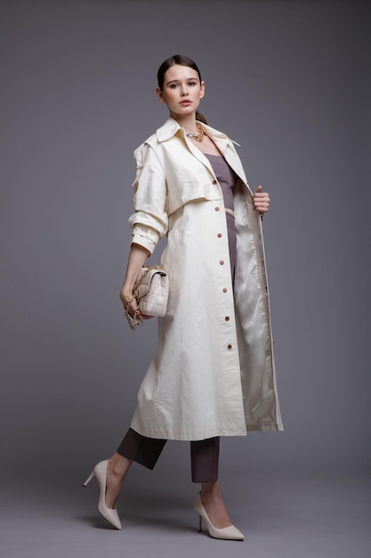 Elegante Frau im hübschen weißen Mantel lila Top Hosen Accessoires Handtasche auf grauem Hintergrund