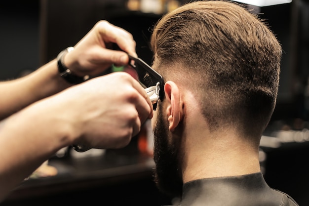 Foto foto de perfil de un joven peluquero recortando el cabello de su cliente con una afeitadora eléctrica y un peine en una barbería.