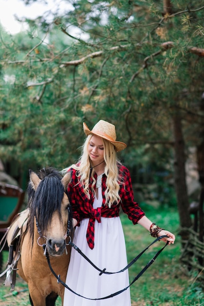 Foto hermosa mujer joven rubia en un elegante sombrero cerca de la vieja valla de madera con ponis país novia
