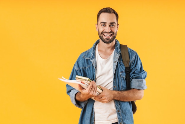 Imagem do aluno de conteúdo em roupas jeans sorrindo enquanto segura cadernos isolados sobre a parede amarela