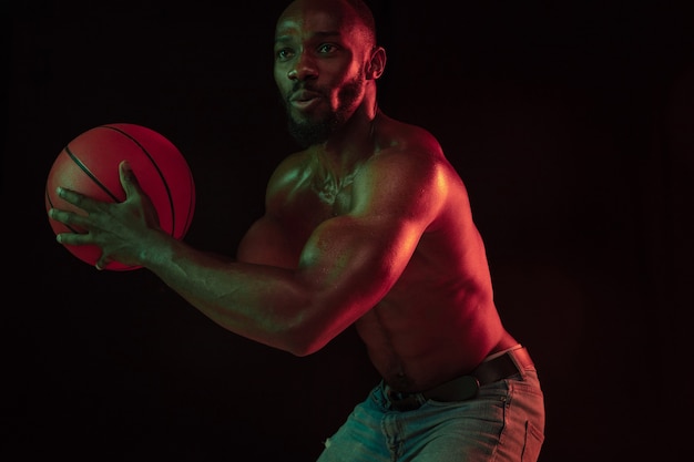 Jogador de basquete jovem musculoso afro-americano em ação de treinamento de jogabilidade, praticando em luzes de néon sobre o fundo escuro do estúdio. Conceito de esporte, movimento, energia, estilo de vida dinâmico e saudável.