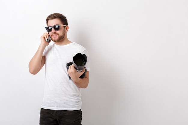 Jovem fotógrafo profissional de camisa branca segurando uma câmera digital pesada com uma lente longa e falando ao telefone com um cliente