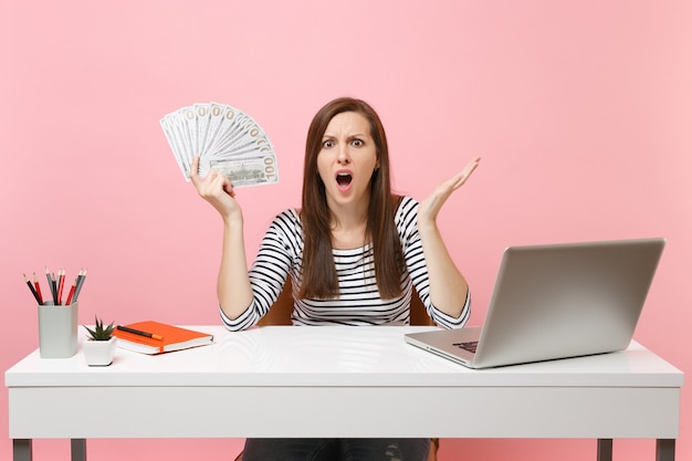 Junge schockierte verwirrte Frau, die Hände mit Bündel vielen Dollar Bargeld ausbreitet, arbeiten im Büro am weißen Schreibtisch mit PC-Laptop