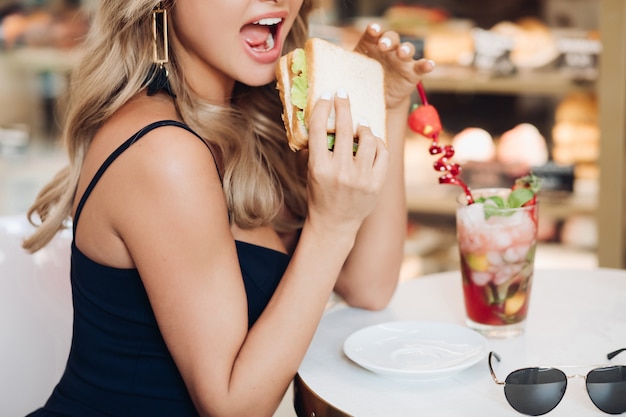 Mittleres Nahaufnahmeporträt der schönen jungen Modefrau, die frisches appetitanregendes Sandwich beißt
