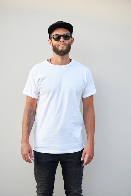 Modelo masculino guapo hipster con barba con camiseta blanca en blanco y una gorra de béisbol con espacio para su logotipo o diseño en estilo urbano casual