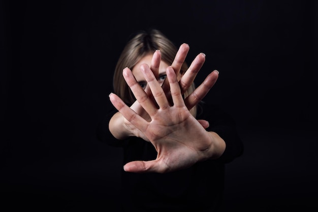 Foto mujer con las manos delante de ella mostrando la señal de stop ocultando la cara en vestido negro sobre fondo negro víctima de abuso físico y psicológico gaslighting agresión relativa