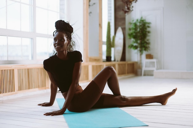 Mulher afro-americana em forma sorridente, virando o corpo sentado no chão com treinamento de perna dobrada no tapete de ginástica em estúdio decorado, alongamento muscular, exercícios ginásticos, fitness, mantendo o corpo em forma