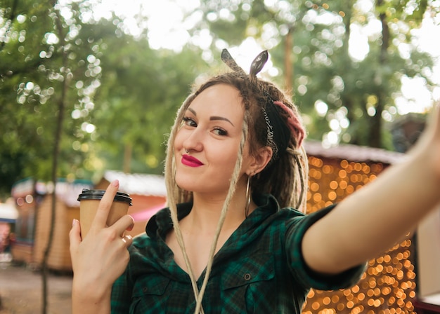 Foto mulher atraente com um corte de cabelo informal dreadlocks faz selfie retrato e segurando a xícara de café