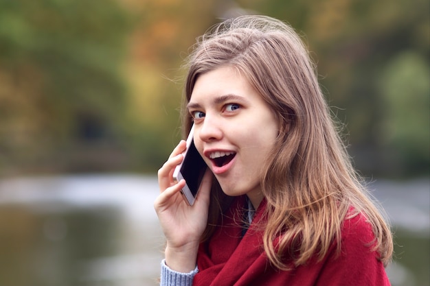 Mulher jovem feliz chocada surpresa falando no celular celular inteligente ao ar livre no parque outono com