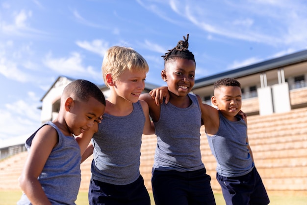 Foto niños de primaria multirraciales sonrientes de pie con el brazo alrededor contra el edificio de la escuela en un día soleado