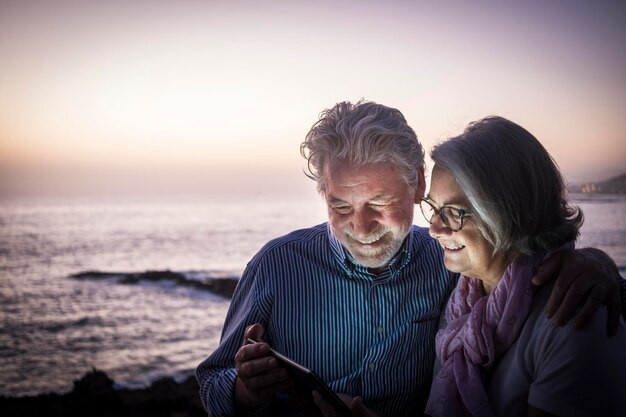 Foto pareja de ancianos mirando una tableta digital por el mar contra el cielo durante la puesta de sol