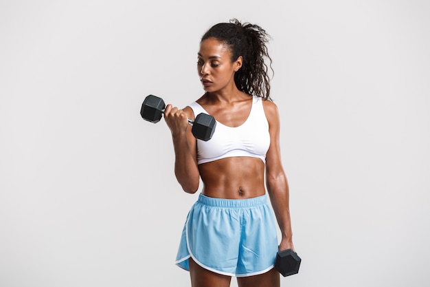 Retrato de uma atraente desportista africana confiante em forma saudável, exercitando-se com halteres isolados sobre uma parede branca