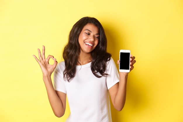 Foto retrato de linda niña afroamericana sonriendo complacido mostrando signo bien y pantalla de teléfono móvil reco ...