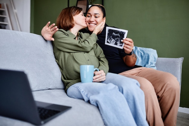 Retrato de una pareja gay besándose esperando un bebé y mostrando una imagen de ultrasonido a la familia a través de un chat de video