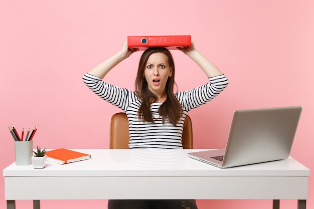 Schockierte Frau, die einen roten Ordner mit Papierdokument auf dem Kopf hält und an einem Projekt arbeitet, während sie im Büro mit Laptop sitzt, isoliert auf pastellrosa Hintergrund. Erfolgsgeschäftskarrierekonzept. Platz kopieren.