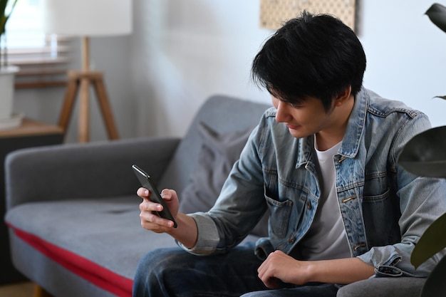 Sorrindo jovem asiático relaxando no sofá em casa e usando telefone celular Conceito de pessoas e tecnologia