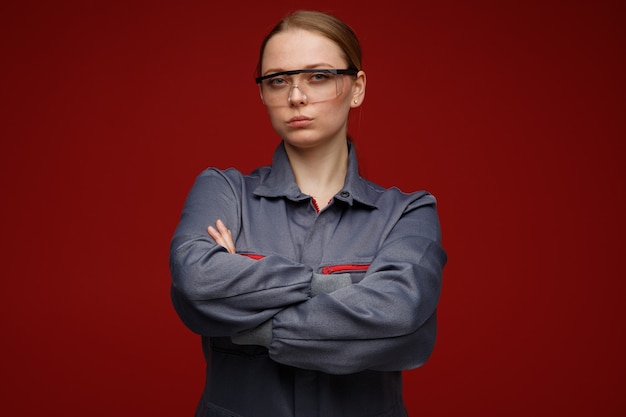 Foto gratuita confiado joven ingeniera rubia vistiendo uniforme y gafas de seguridad de pie con postura cerrada