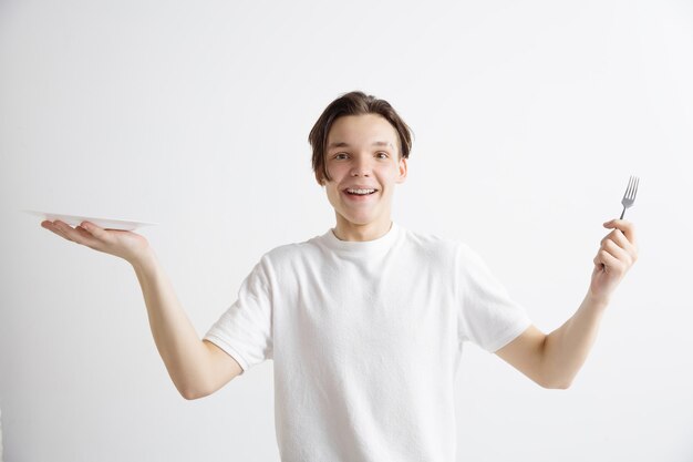 Foto gratuita chico caucásico atractivo sonriente joven con plato vacío y tenedor aislado sobre fondo gris. copie el espacio y simule. fondo de plantilla en blanco.