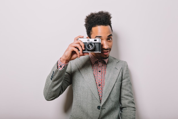 Foto gratuita chico americano-africano con estilo moderno posando en la pared blanca con una cámara retro y toma disparos en gris