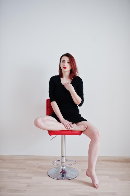 Foto gratuita chica pelirroja con túnica negra sentada en una silla roja contra una pared blanca en una habitación vacía