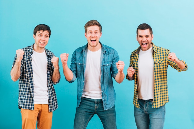Foto gratuita amigos masculinos jovenes emocionados que aprietan su puño contra fondo azul