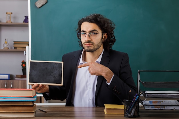 Foto gratuita estricto profesor con gafas sosteniendo y apunta a la mini pizarra sentado a la mesa con herramientas escolares en el aula