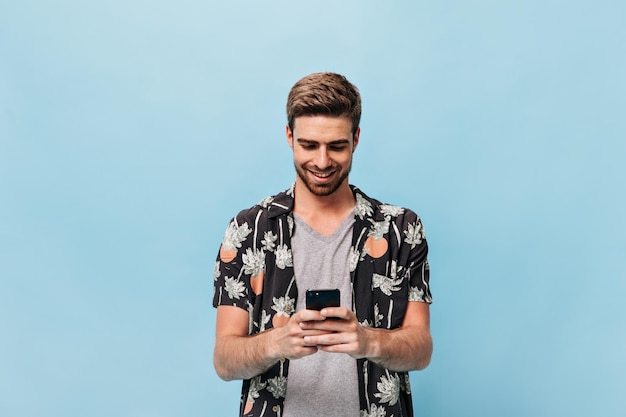 Foto gratuita hombre atractivo con peinado moderno y barba de jengibre en camisa con estampado de piña y palma y camiseta gris sonriendo y sosteniendo el teléfono