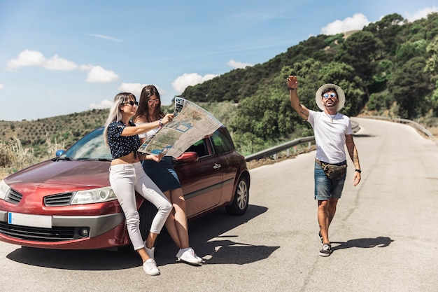 Foto gratuita hembras mirando el mapa apoyado en el coche mientras su amigo masculino gesticulando en la carretera