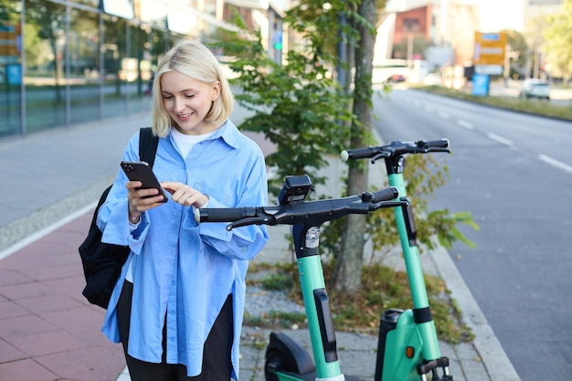 Foto gratuita imagen de una joven sonriente que se dirige al trabajo alquilando usando una aplicación de teléfono móvil para desbloquear un scooter callejero