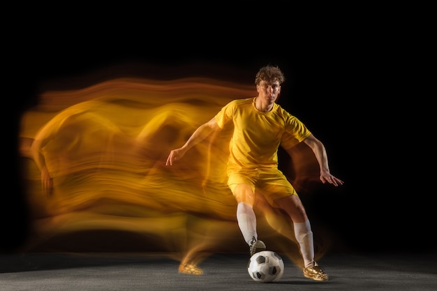 Foto gratuita jugador de fútbol o fútbol masculino caucásico joven pateando la pelota para el gol en luz mixta en la pared oscura concepto de pasatiempo deportivo profesional de estilo de vida saludable