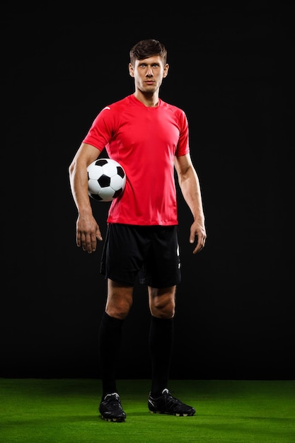 Foto gratuita jugador de fútbol en uniforme negro y rojo con balón en la mano de pie sobre el césped sobre la pared negra, mirando al frente
