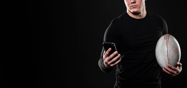 Foto gratuita jugador de rugby masculino con smartphone mientras sostiene la pelota con espacio de copia