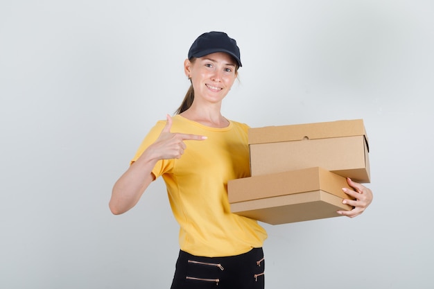 Foto gratuita mujer de entrega en camiseta, pantalón, gorra apuntando a cajas de cartón y mirando alegre