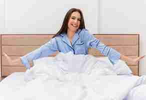Foto gratuita mujer hermosa joven en pijama azul sentada en la cama descansando sobre suaves almohadas despertando feliz y positivo sonriendo disfrutando el fin de semana en el interior del dormitorio