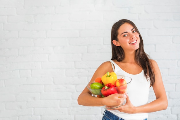 Foto gratuita mujer joven atractiva de pie contra la pared sosteniendo un tazón de verduras frescas y frutas