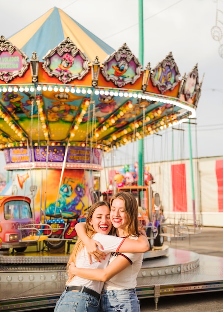 Foto gratuita mujer joven sonriente dos que abraza delante del carrusel colorido iluminado